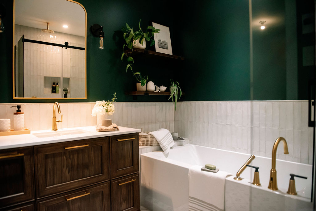 Salle de bain élégante avec murs verts, meuble-lavabo en bois, luminaires dorés et baignoire blanche. Des étagères accueillent plantes et décorations. Un grand miroir est suspendu au-dessus du lavabo.