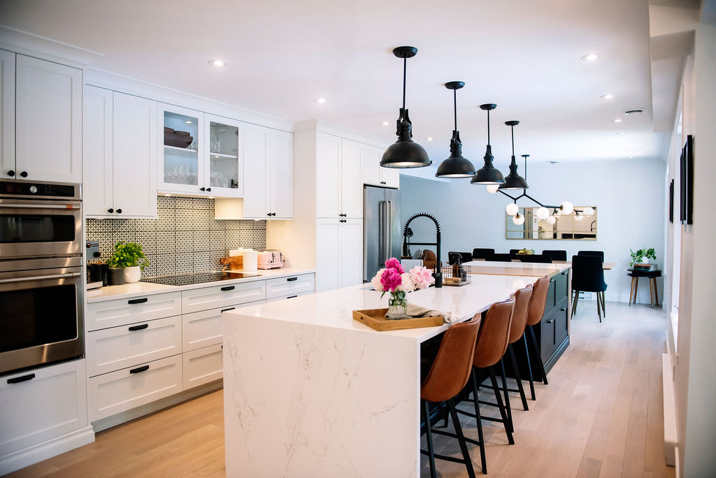 Cuisine moderne avec armoires blanches, grand îlot en marbre, tabourets en cuir marron, luminaires suspendus et planchers de bois franc. Un coin repas est visible en arrière-plan.