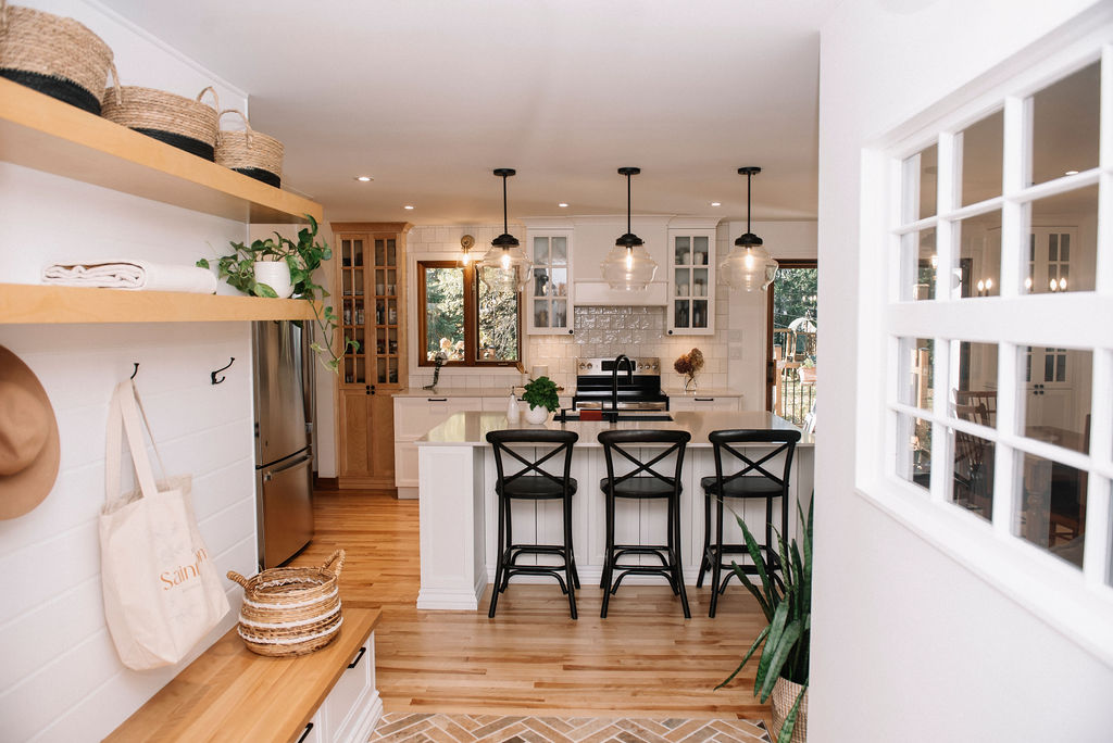 Cuisine moderne avec parquet, îlot central avec tabourets de bar, luminaires suspendus et étagères adjacentes. Des plantes vertes et des paniers tressés ajoutent des touches décoratives.