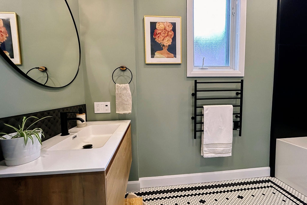 Une salle de bain moderne avec une coiffeuse en bois, un miroir rond, un porte-serviettes, des œuvres d'art murales et une plante. Le sol est en carrelage hexagonal noir et blanc et une fenêtre givrée apporte de la lumière naturelle.